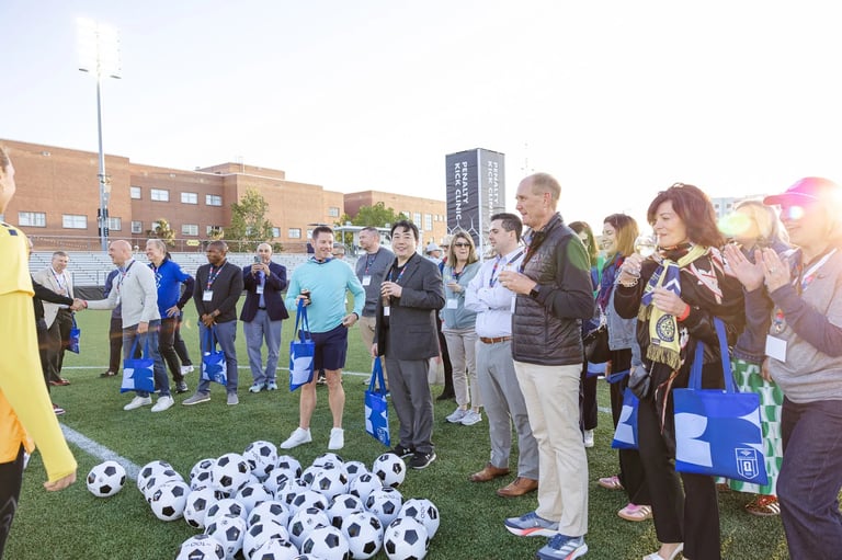 Group of people gathered on grass field with soccer balls during outdoor community event near brick buildings