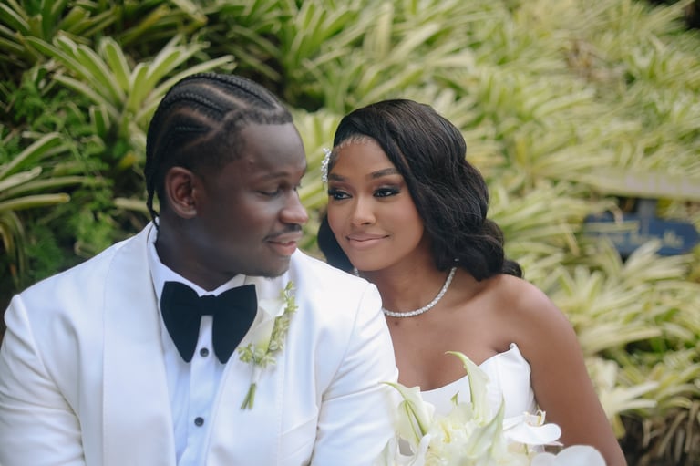 A groom and bride in wedding attire pose together outdoors with green foliage in the background
