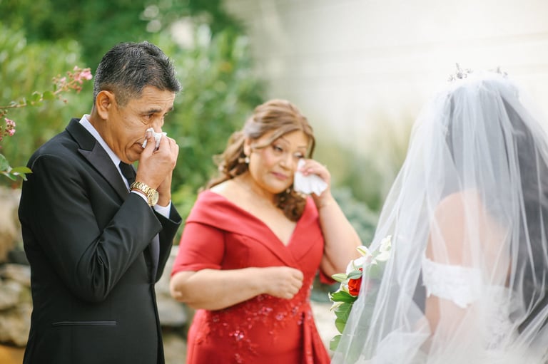 Emotional father in black suit wipes tears while bride in white veil stands nearby at outdoor wedding ceremony