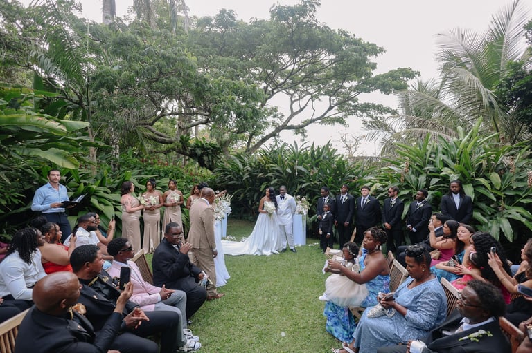 Outdoor garden wedding ceremony with guests seated on grass, bride and groom in center, surrounded by tropical plants and trees