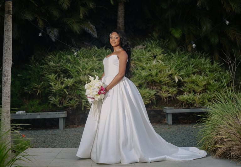 Bride in white sleeveless wedding gown with flowing train holding bouquet in tropical garden setting at night