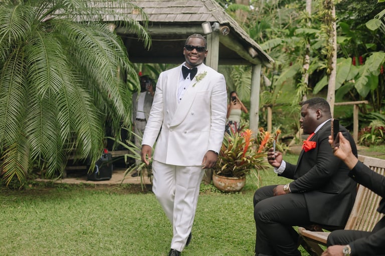 Man in white suit and sunglasses walking on grass while person in black suit photographs him in tropical garden setting