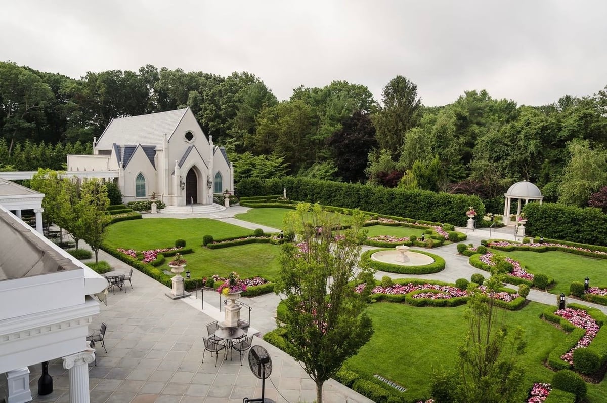 White chapel with arched windows set in manicured gardens featuring green lawns, flower beds, and tree-lined pathways on a cloudy day
