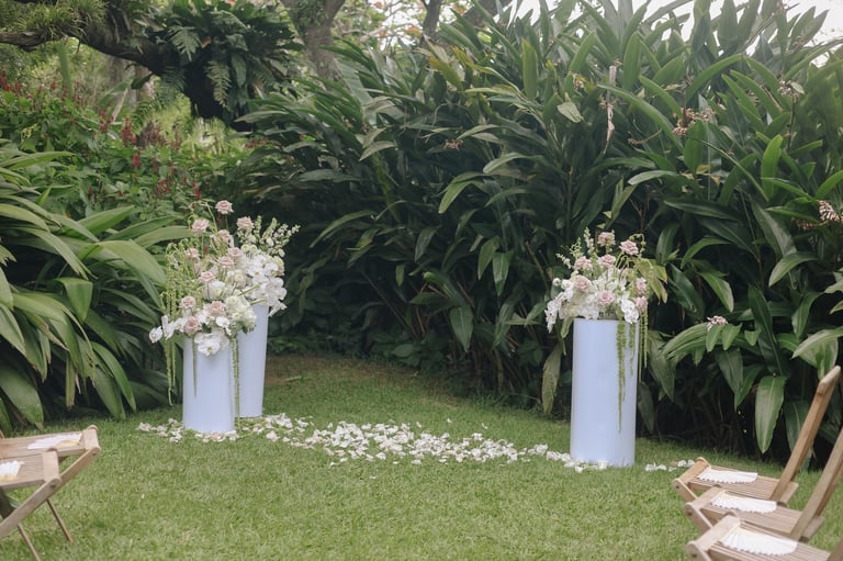 Garden wedding ceremony setup with white floral pedestals, green lawn, and lush tropical plants forming a natural backdrop