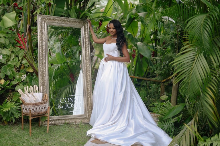 Woman in white flowing dress posing near ornate mirror surrounded by tropical plants in lush garden setting