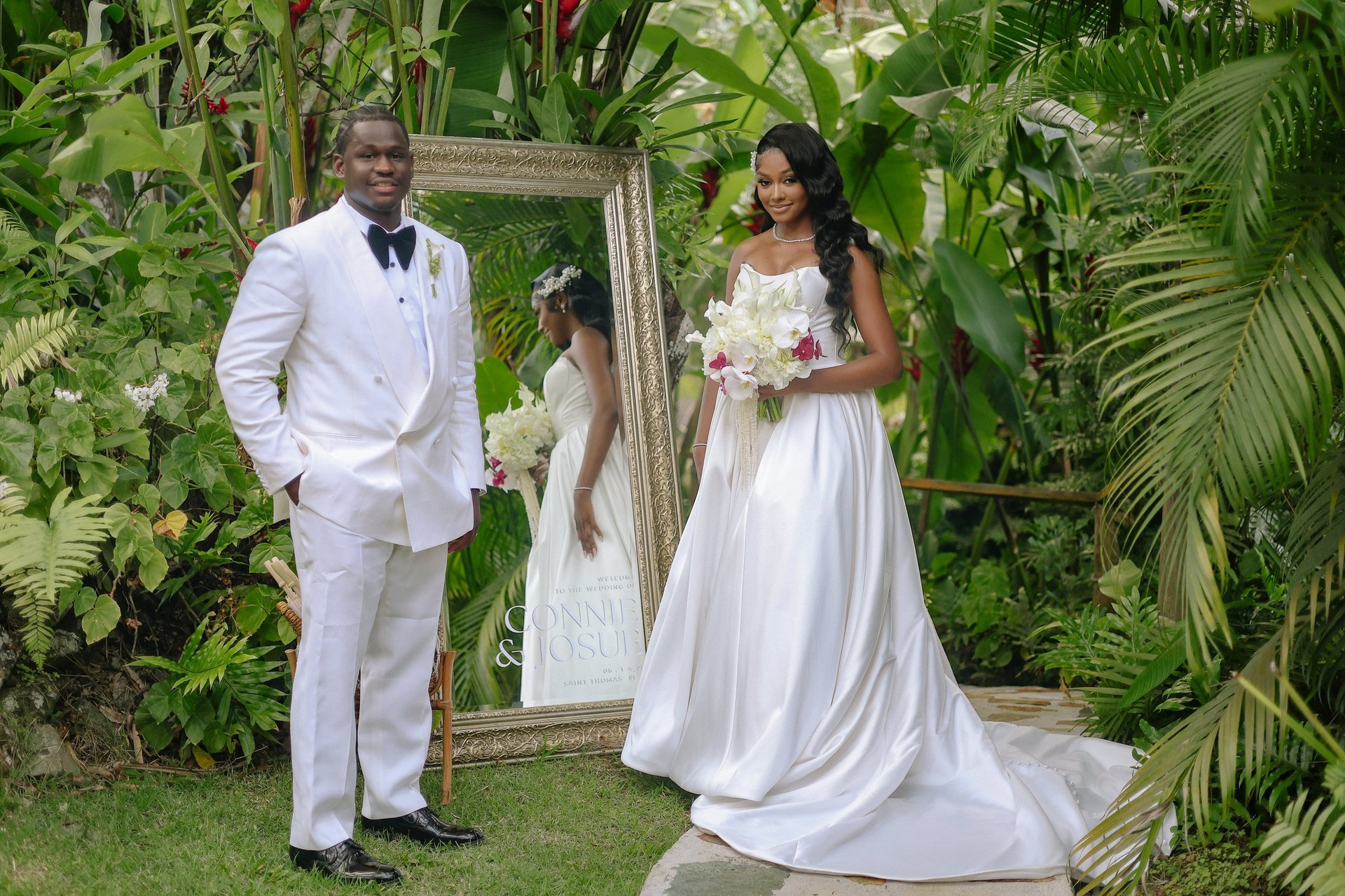 Groom in white suit standing with bride in white wedding dress holding bouquet in front of wooden mirror frame in tropical garden setting