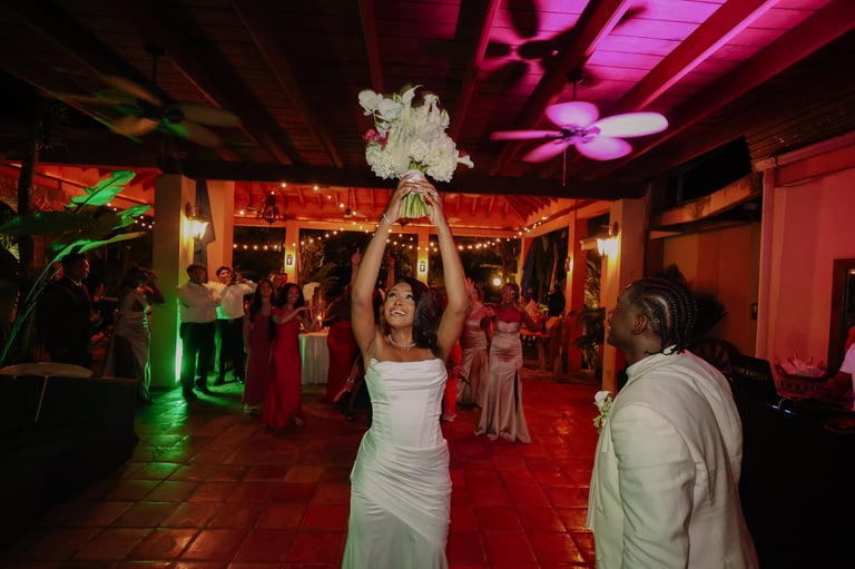 Bride in white dress tosses bouquet at wedding reception with guests under pink and red ambient lighting