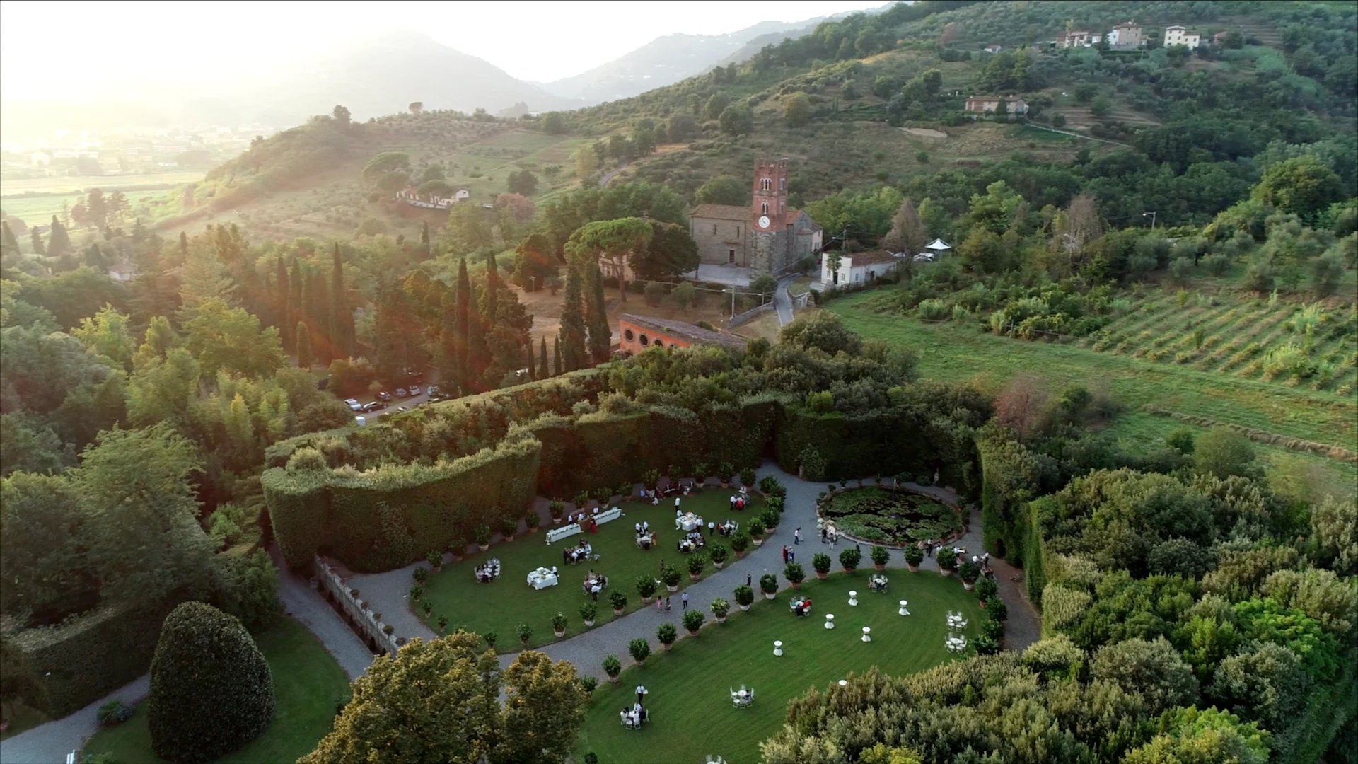 Aerial view of a Tuscan villa with manicured gardens, swimming pool, and cypress trees surrounded by rolling hills at sunset