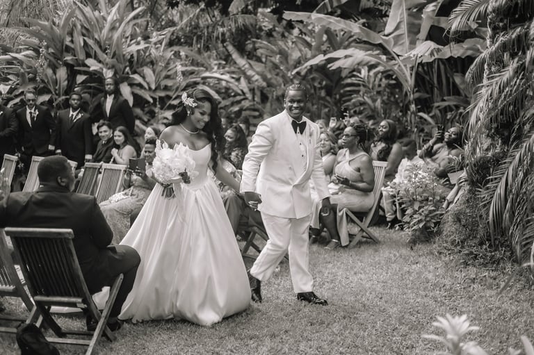 Black and white photo of bride and groom walking together in tropical garden wedding ceremony with guests seated on either side