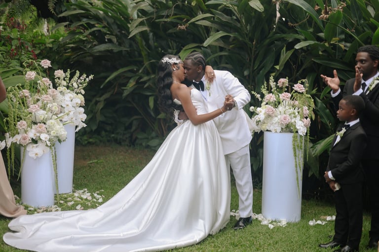 Bride and groom kissing during outdoor wedding ceremony with white floral arrangements and green foliage backdrop