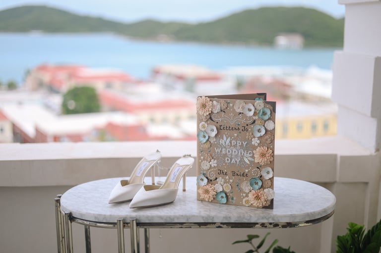 Wedding day book and white bridal heels displayed on a round table with a waterfront harbor view in the background
