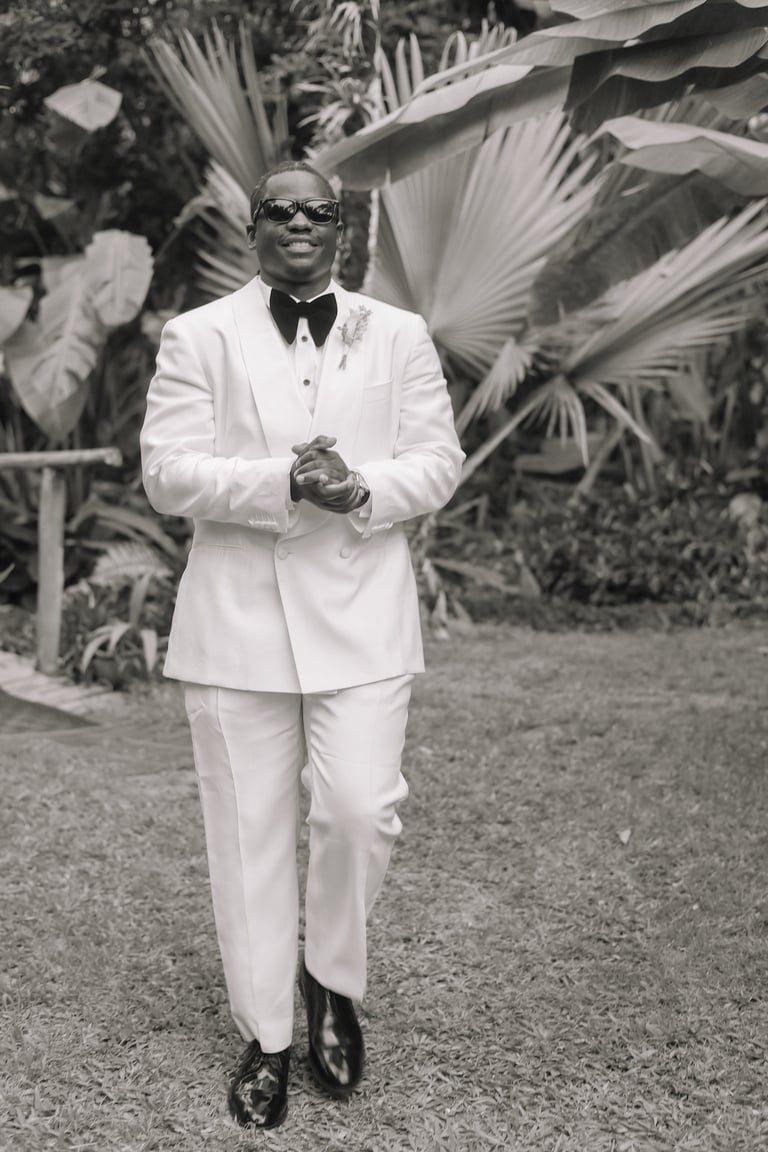 Man in white tuxedo and sunglasses walking outdoors with tropical palm foliage in background, black and white photograph