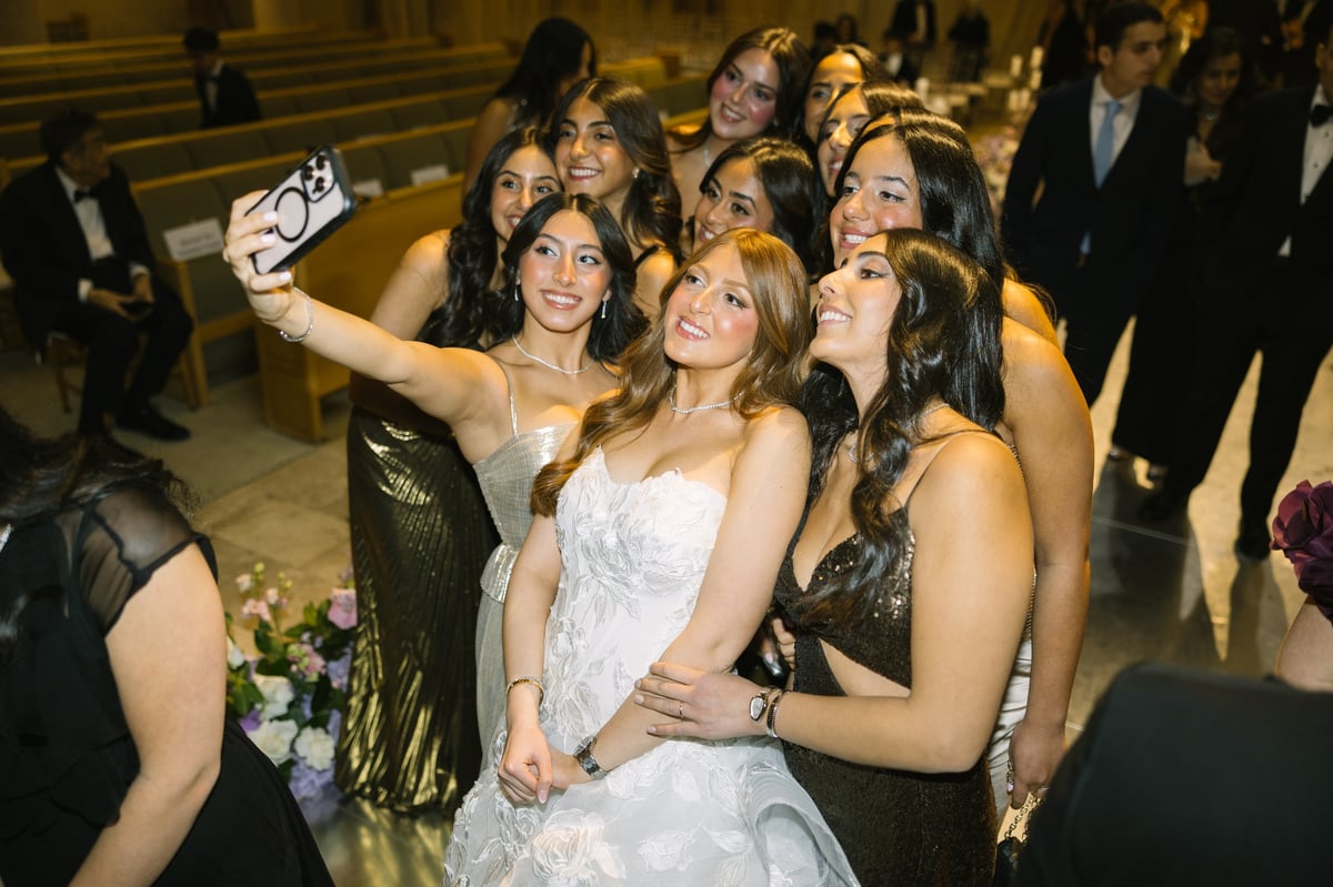 Bride in white dress taking a selfie with female friends at a formal event, indoors at what appears to be a wedding reception or celebration
