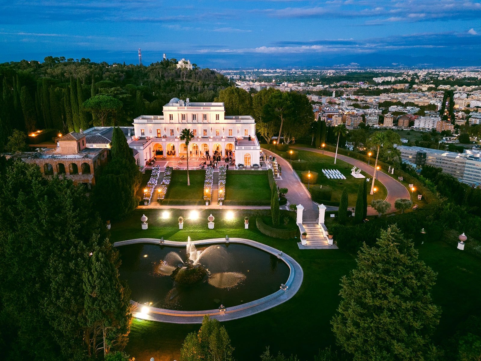 Aerial view of illuminated pink villa with manicured gardens, circular pond, cypress trees, and city skyline at dusk
