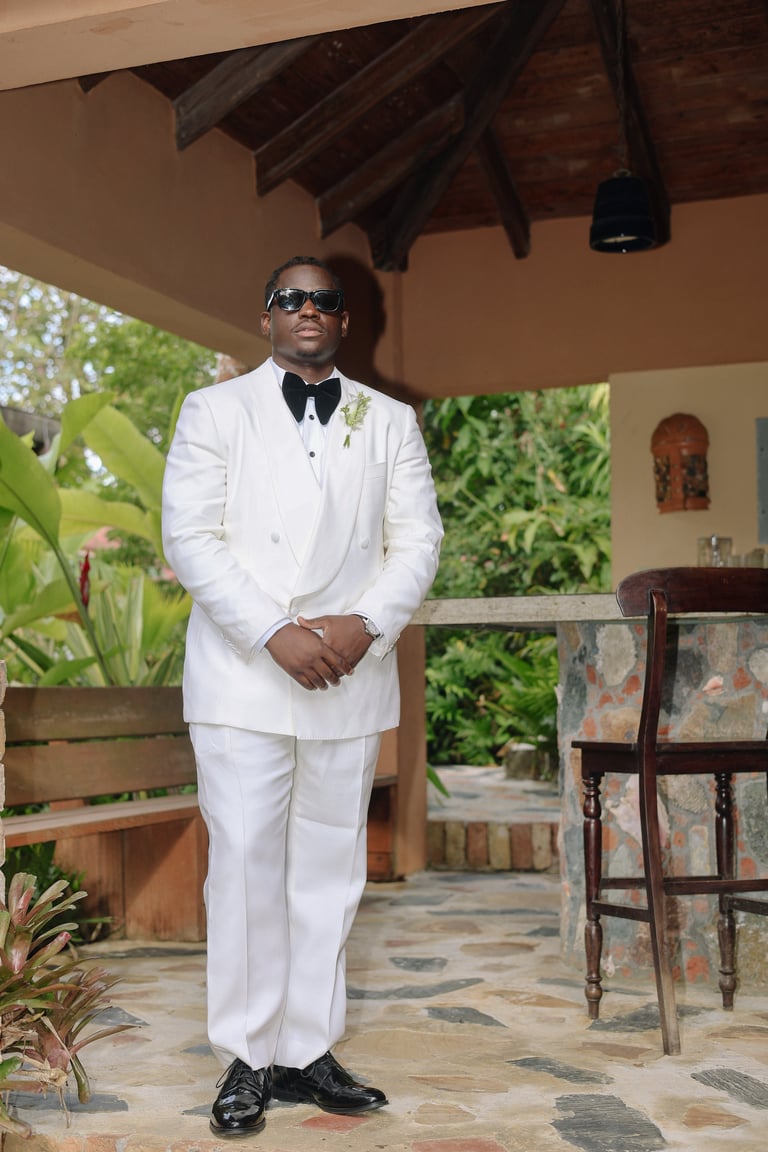 Man in white double-breasted suit and sunglasses standing under a covered patio with tropical plants and wooden furnishings