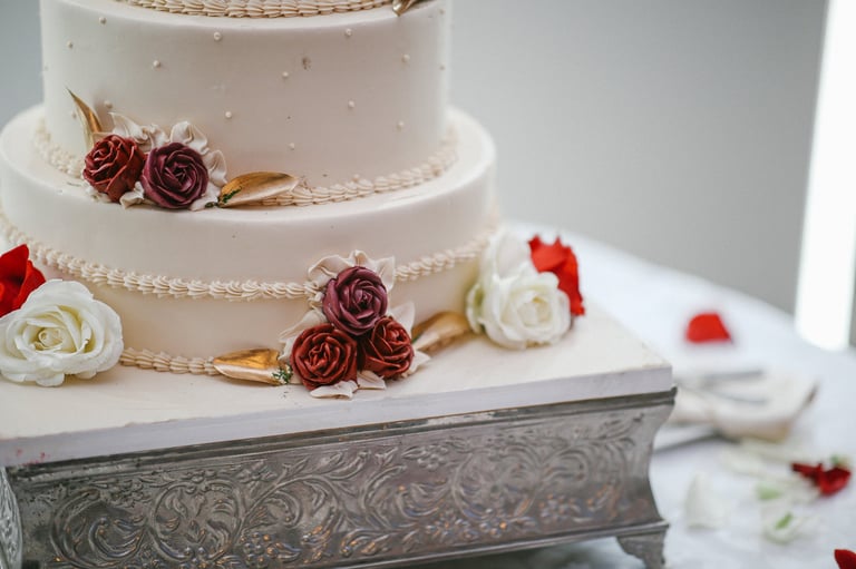 Three-tiered white wedding cake with burgundy and cream roses, gold ribbons, and pearl beading on an ornate silver cake stand