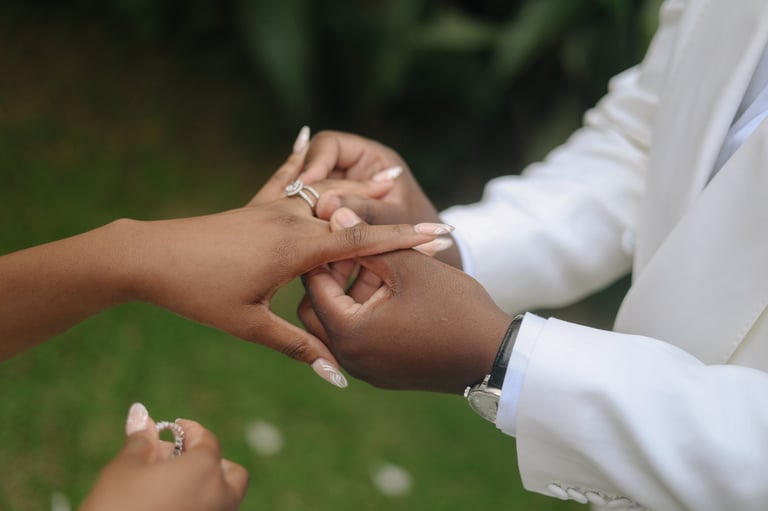 Close-up of bride and groom hands exchanging wedding rings, wearing white formal attire with green background
