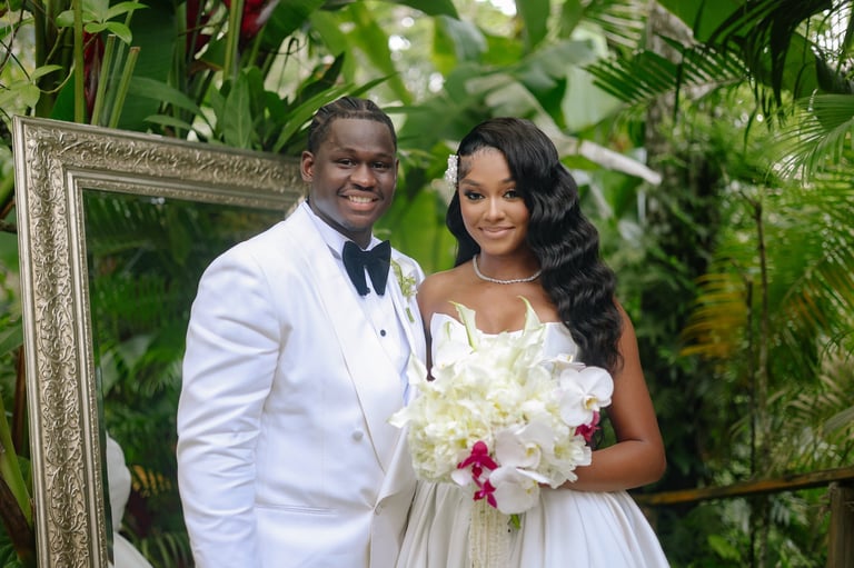 A groom in a white tuxedo and bow tie stands with a bride in a white dress holding a white and pink bouquet in a lush garden setting with a decorative frame behind them
