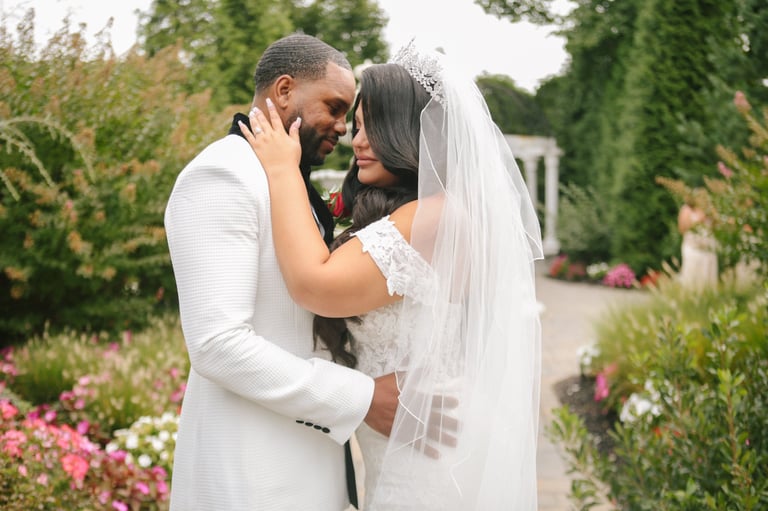 Bride and groom embrace and kiss in a lush garden setting with pink flowers and green hedges