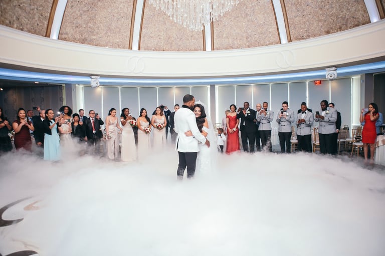 Wedding couple dancing on a dance floor filled with dry ice fog, surrounded by guests in formal attire in a curved hall with skylights