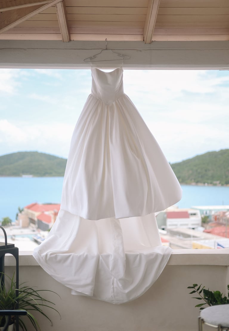 White wedding dress hanging on wooden beam near window overlooking coastal landscape with mountains and blue water
